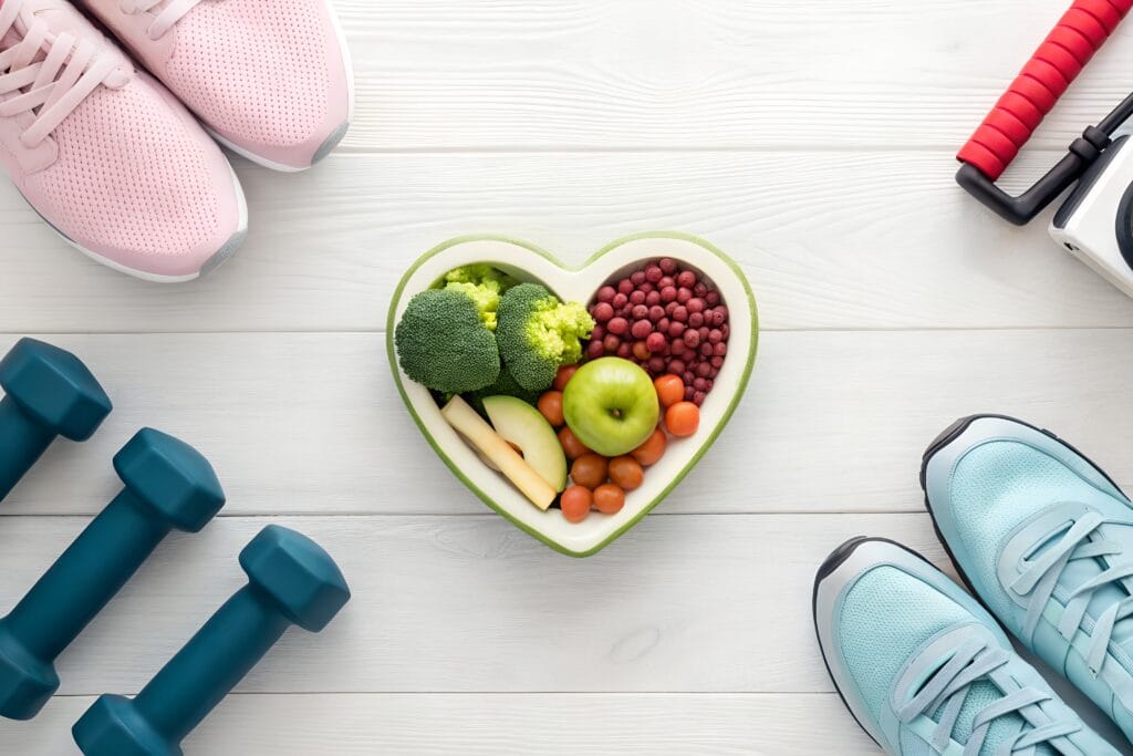 وعاء على شكل قلب يحتوي على منتجات طازجة محاطة بأحذية رياضية وأثقال على خلفية خشبية بيضاء.